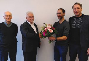 Ein Mann übergibt einem anderen Mann einen Blumenstrauß. Rechts und links davon stehen zwei weitere Männer. Sie stehen vor einer hellen Wand und blicken in die Kamera.