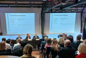 Siezendes Publikum von hinten zu sehen, blickt auf das Podium mit vier Personen, an der Wand zwei Bildprojektionen