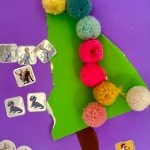 Lilafarbener Untergrund, grünes Dreieck, brauner Stamm, darüber und daneben kleine farbige Wollbommeln und kleine glitzernde Aufkleber