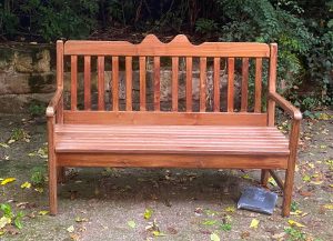 Eine hölzerne Bank steht auf einem Steinboden im Freien, Blätter liegen herum, im Hintergrund eine niedrige Mauer und Büsche. Unter der Bank liegt ein flaches graues Päckchen, offenbar eine Abdeckplane.