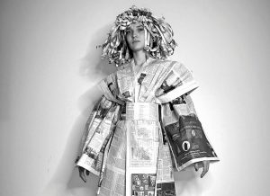 Schwarzweiß-Foto: Person in Kleidung und mit Perücke aus Zeitungspapier vor heller Wand