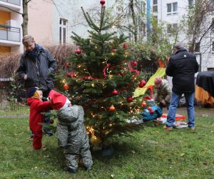 Geschmückter Weihnachtsbaum auf einer Wiese, zwei Kinder im Vordergrund, weitere Personen im Hintergrund