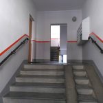 Breiter Treppenaufgang, Stufen, schwarze Handläufe an beiden Seiten, Briefkästen; Wände weiß und grau mit rotem Streifen.