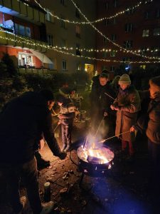 Menschen stehen im weihnachtlich geschmückten Hof um eine Feuerschale und halten lange Stöcker mit Teig in die Flammen